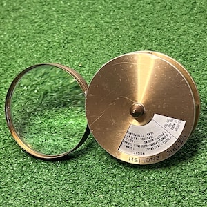 May include: A circular brass conversion tool featuring a clear glass lens on one side and a metal disc on the other. The disc displays printed text for metric and English weight and length conversions, including values like 1 ounce equals 28.35 grams.