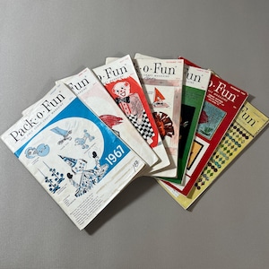 May include: A collection of vintage "Pack-o-Fun" magazines from 1967, featuring colorful covers with illustrations of clowns, sailboats, and other craft-related designs. The magazines are stacked, showcasing various issues.