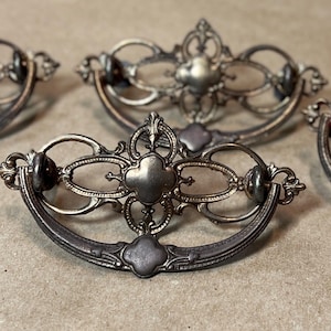 May include: Antique silver-toned drawer pulls with ornate, symmetrical designs. Each pull features a curved handle and decorative centerpieces with floral and geometric patterns. The hardware is likely made of metal and is suitable for furniture restoration or crafting projects.