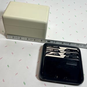 May include: A cream-colored, rectangular address book box with a lid, next to a black address book with labeled tabs. A measuring tape shows the box is approximately 5 inches long.