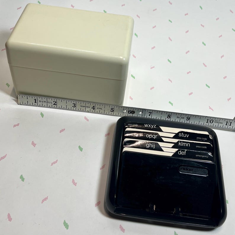 May include: A cream-colored, rectangular address book box with a lid, next to a black address book with labeled tabs. A measuring tape shows the box is approximately 5 inches long.