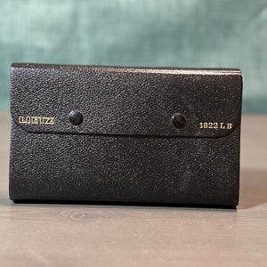 May include: A black, textured case with a snap closure. The case has the text "LILITZ" and "1822 LB" in gold lettering. The case is rectangular and appears to be made of leather or a similar material.