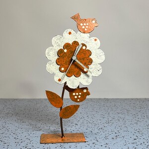 May include: A metal table clock shaped like a flower with white and orange petals. The center is a dark orange flower head with clock hands. Two small orange birds with white polka dots are attached to the top and bottom. It has a thin stem and a rectangular base.