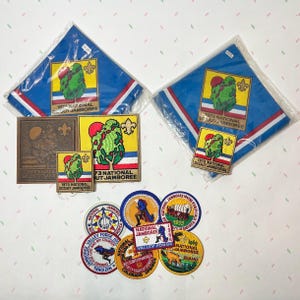 May include: Collection of vintage Boy Scout memorabilia, including blue neckerchiefs, patches, and a metal plaque. The items feature the text "1973 National Scout Jamboree" and other event-related designs. Various patches showcase different locations and years.