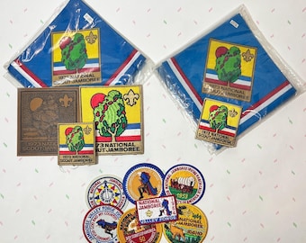 Puede incluir: Colección de recuerdos vintage de los Boy Scouts, que incluyen pañuelos azules, parches y una placa de metal. Los artículos presentan el texto "1973 National Scout Jamboree" y otros diseños relacionados con el evento. Varios parches muestran diferentes ubicaciones y años.
