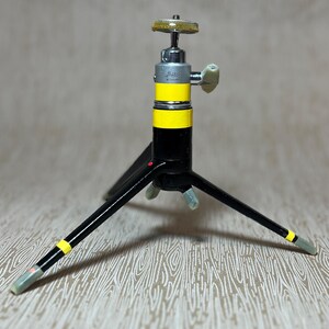 May include: A vintage black and yellow tripod with three legs. The tripod has a silver adjustable head with a yellow and black striped center section. The legs have yellow stripes near the feet.