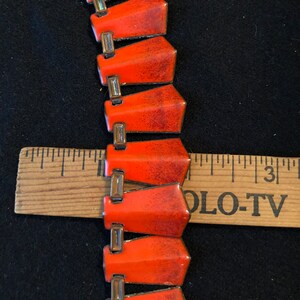 May include: A close-up of a red and gold enamel necklace. The necklace is made of a series of connected, pointed, red enamel pieces with gold accents. The necklace is lying on a black surface with a wooden ruler beside it.