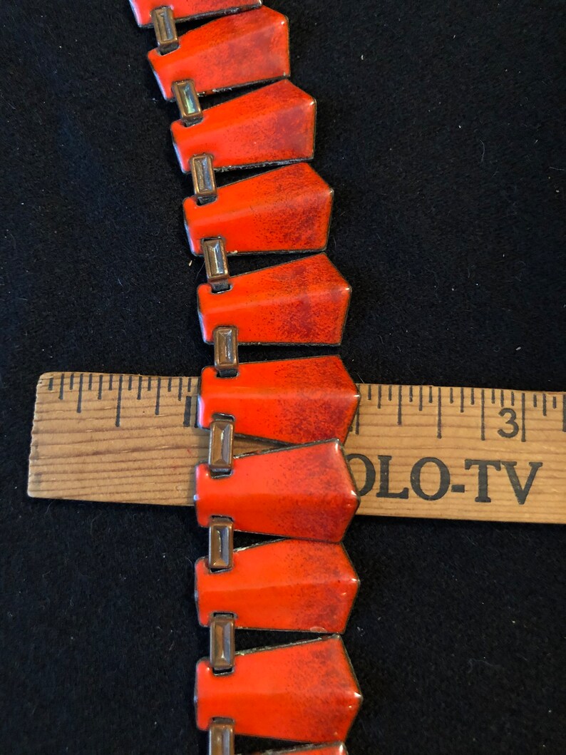 May include: A close-up of a red and gold enamel necklace. The necklace is made of a series of connected, pointed, red enamel pieces with gold accents. The necklace is lying on a black surface with a wooden ruler beside it.
