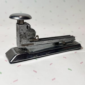 May include: A vintage, chrome-plated stapler with a rounded top and a lever. The stapler shows signs of wear, with a slightly tarnished finish. The base is rectangular, and the overall design is classic and functional.