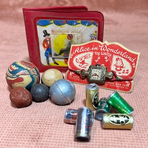 May include: A collection of vintage toys including a red and white toy watch with a white rabbit and the words "Alice in Wonderland Toy Watch" on the face, a red and white toy wallet with a picture of a clown and a dog, a red and white rubber ball, a white and black rubber ball, a silver metal ball, a brown and black stone, a red stone, and five small silver cans with various designs.