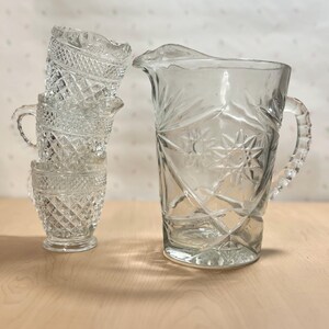 May include: A clear glass pitcher with a starburst design and a handle, next to a stack of three small, matching glass cups. The cups and pitcher have a diamond pattern.
