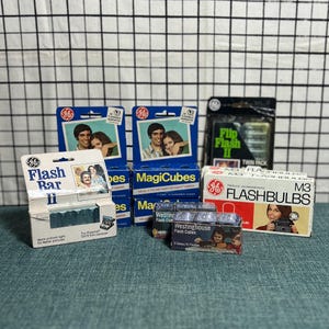 May include: A collection of vintage camera flash accessories, including GE Flash Bar II, Magicubes, Flip Flash II, and M3 Flashbulbs. The packaging features retro graphics and text, with images of people and cameras. The items are arranged on a teal surface.
