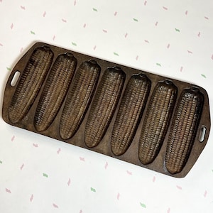 May include: A cast iron cornbread pan with seven corn cob molds. The pan is dark brown with handles on each end. The molds are detailed to resemble corn cobs.