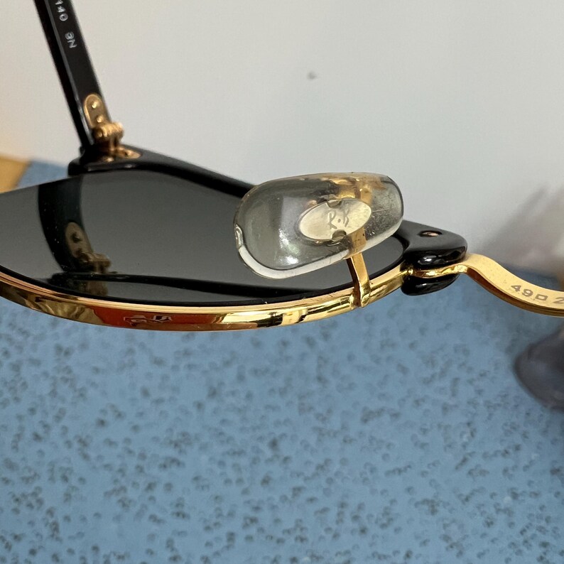 Op de afbeelding: Close-up van een zonnebril met zwarte glazen en goudkleurige monturen. De neuspads zijn transparant en de pootjes zijn zwart. De zonnebril heeft een klassiek design.