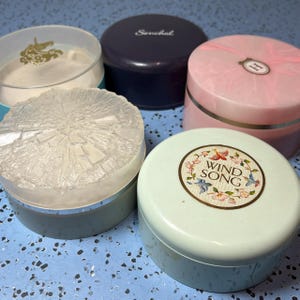 May include: An assortment of vintage cosmetic containers. The containers are in various colors, including light blue, pink, and navy. One container has the text "Wind Song" in a floral design. Another has the text "Senchal".