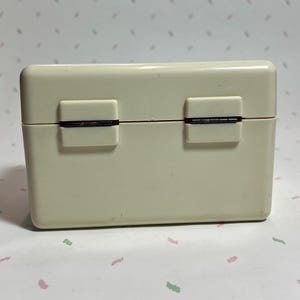 May include: A rectangular, off-white plastic box with a hinged lid. The box has two rectangular clasps on the front and a visible hinge. The box is likely a container for small items.