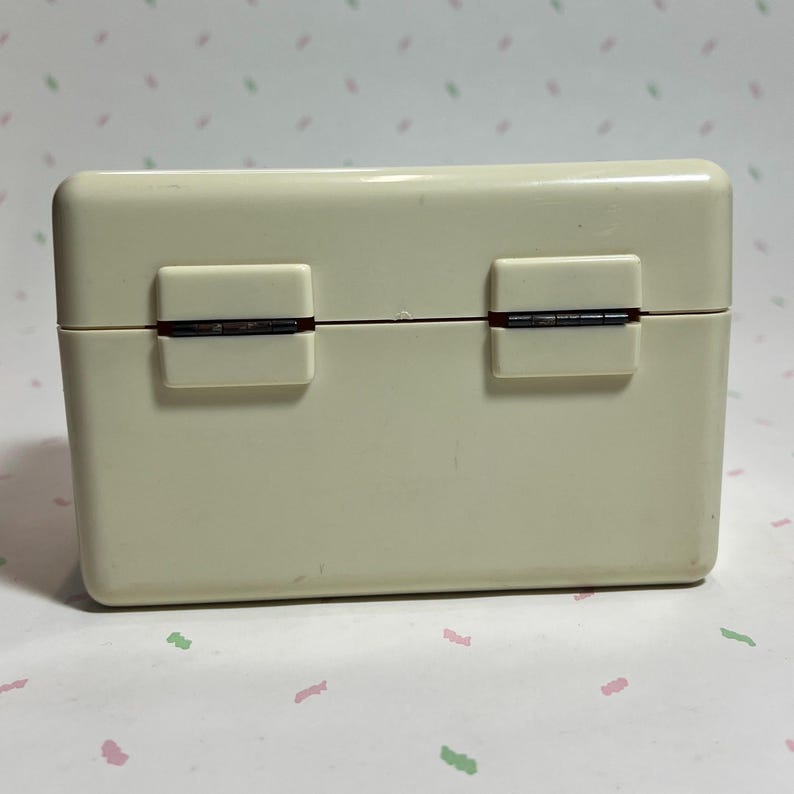 May include: A rectangular, off-white plastic box with a hinged lid. The box has two rectangular clasps on the front and a visible hinge. The box is likely a container for small items.
