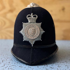 May include: A black police helmet with a silver emblem featuring a crown and the words "Essex Constabulary." The helmet has a rounded top and a curved brim. The emblem is a starburst shape with the words "Essex Constabulary" around the edge.