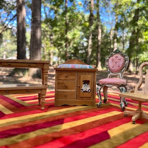 May include: A set of miniature wooden furniture, including a changing table with a teddy bear design, a small table with two shelves, a pink and white chair with a star design, and a wooden chair with a high back. The furniture is arranged on a striped rug.