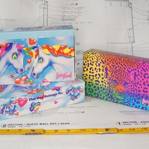 May include: Two rectangular cardboard boxes with Lisa Frank branding. One box features two white unicorns with rainbow manes and hearts against a blue sky. The other box has a vibrant rainbow leopard print. A yellow ruler is visible at the bottom.