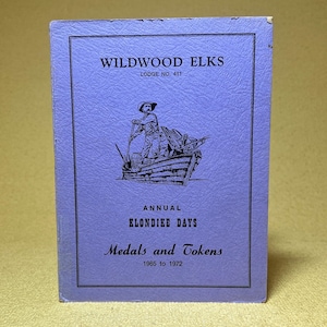 May include: A vintage, lavender-colored booklet titled "Wildwood Elks Lodge No. 411." The cover features a black and white illustration of a man in a boat. Text includes "Annual Klondike Days" and "Medals and Tokens 1965 to 1972."