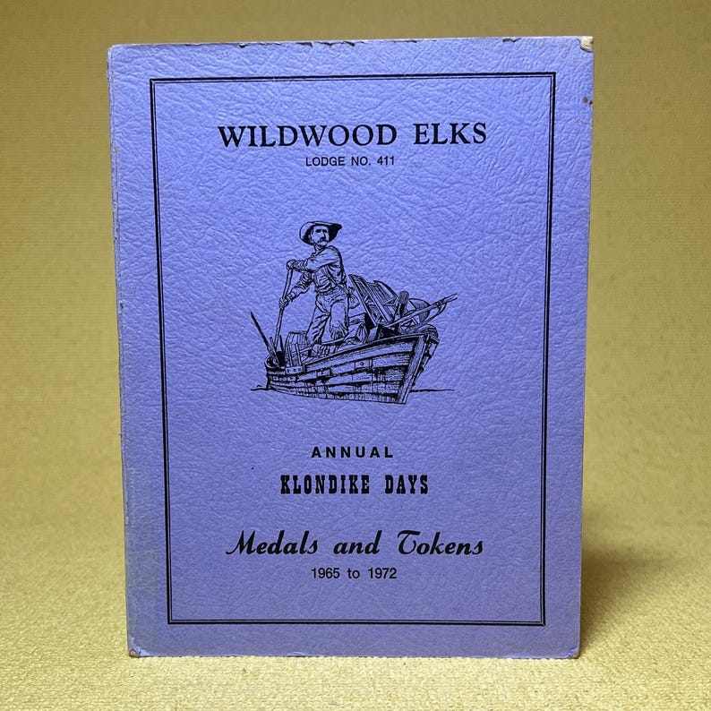May include: A vintage, lavender-colored booklet titled "Wildwood Elks Lodge No. 411." The cover features a black and white illustration of a man in a boat. Text includes "Annual Klondike Days" and "Medals and Tokens 1965 to 1972."