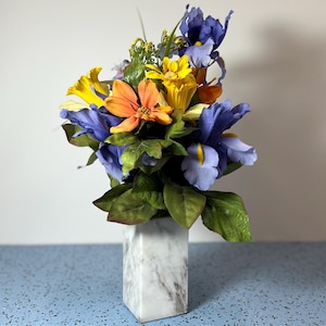 May include: A floral arrangement featuring blue irises, yellow and orange flowers, and green leaves. The flowers are arranged in a white marble square vase. The background is a light blue surface and a white wall.