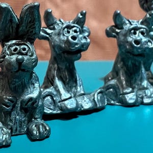 May include: A set of four pewter figurines, including a rabbit and three bulls. The figurines are all in a sitting position and have a whimsical, cartoonish style. They are painted in a dark gray color.