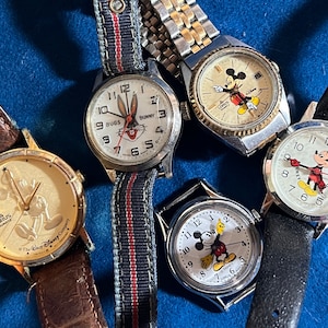 May include: Five wristwatches with different cartoon characters on the face. The watches have various colored bands, including brown leather, black leather, and a blue and red striped band. The characters include Mickey Mouse, Bugs Bunny, and a rabbit.