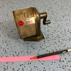 May include: An antique pencil sharpener with a metal body and a crank handle. The sharpener has a label that reads "APSCO CHICAGO". A black pencil rests on a pink strip of paper.