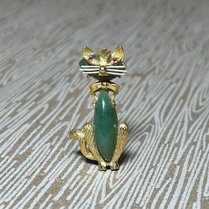 May include: Gold-toned cat brooch with a green jade body and pink gemstone eyes. The cat's head features gold ears and white whiskers. The brooch is a vintage jewelry piece.