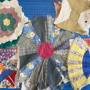 May include: A collection of colorful, vintage fabric pieces arranged on a blue cutting mat. The pieces include floral, striped, and patterned fabrics in various shapes, including a central flower design with a pink center and blue petals.