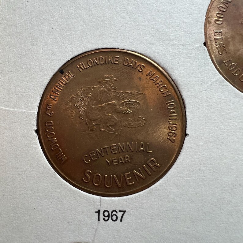 May include: A copper-colored souvenir coin from the 4th Annual Wildwood Klondike Days, March 10-11, 1967. The coin features an image of a person with a horse and a cart, with the words "Centennial Year Souvenir."