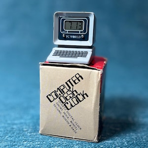May include: A miniature computer desk clock with a digital display showing 7:13. The clock is gray and sits atop a cardboard box with the words "COMPUTER DESK CLOCK" printed on it. The box also states it shows the hour, minute, second, month, and date.