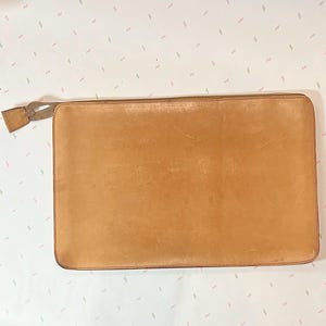 May include: A rectangular, light brown leather clutch with a zipper closure. The clutch has a small leather tab attached to the zipper pull. The leather has a slightly worn appearance.