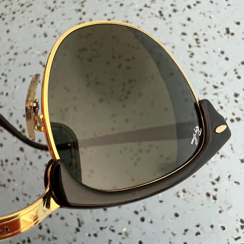 Op de afbeelding: Close-up van een Ray-Ban zonnebril met goudkleurige montuur en donkere glazen. De veren zijn zwart met een gouden accent. Het Ray-Ban logo is zichtbaar op de veer. Deze zonnebril is ontworpen om de ogen te beschermen tegen de zon.