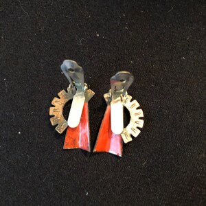 May include: A pair of red and silver clip-on earrings with a geometric design. The earrings have a silver gear-like design on the back and a red triangle shape on the front.
