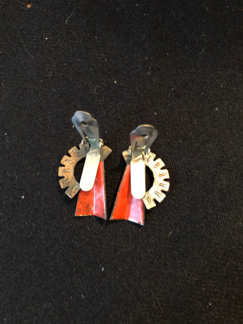 May include: A pair of red and silver clip-on earrings with a geometric design. The earrings have a silver gear-like design on the back and a red triangle shape on the front.