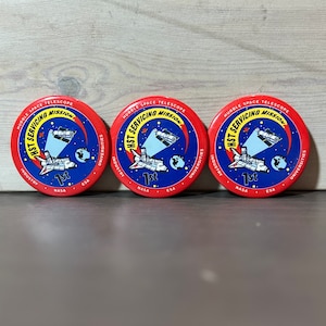 May include: Three red and blue round buttons with the text "Hubble Space Telescope HST Servicing Mission 1st" and the logos of NASA, ESA, Industry and Universities.