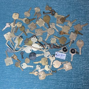 May include: A collection of vintage keys in various shapes and sizes, made of brass and other metals. Some keys have decorative designs, while others have stamped numbers or text. The keys are scattered on a blue textured surface.