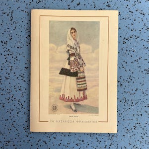 May include: A vintage postcard featuring a woman in traditional Greek attire. The illustration shows a woman in a white dress, a patterned shawl, and a head covering. The postcard has text in Greek.