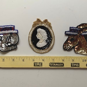 May include: Three beaded patches, one with a silver elephant wearing a blue and white hat with the word "VOTE" on it, one with a white silhouette of a woman in a gold frame, and one with a gold donkey wearing a blue and white hat.