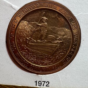 May include: A bronze-colored commemorative coin for the Wildwood 9th Annual Klondike Days, March 3 & 4, 1972. The coin features a man in a boat with a pole, with text around the edge. The text includes "Wildwood Community Hall" and "Good for $1.00 at Wildwood".