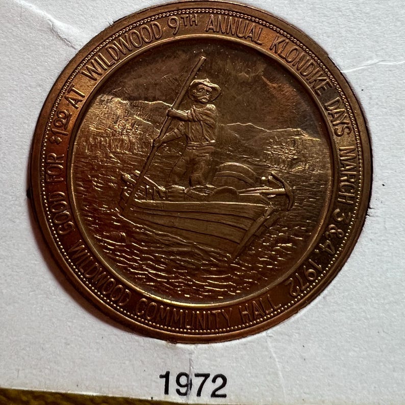 May include: A bronze-colored commemorative coin for the Wildwood 9th Annual Klondike Days, March 3 & 4, 1972. The coin features a man in a boat with a pole, with text around the edge. The text includes "Wildwood Community Hall" and "Good for $1.00 at Wildwood".
