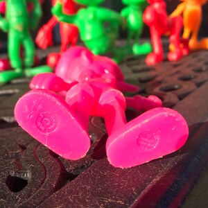 May include: A pink plastic toy figure of a person lying on its back. The figure has a simple design with no facial features. The figure is made of a bright pink plastic and has a smooth surface. The figure is lying on a black surface.