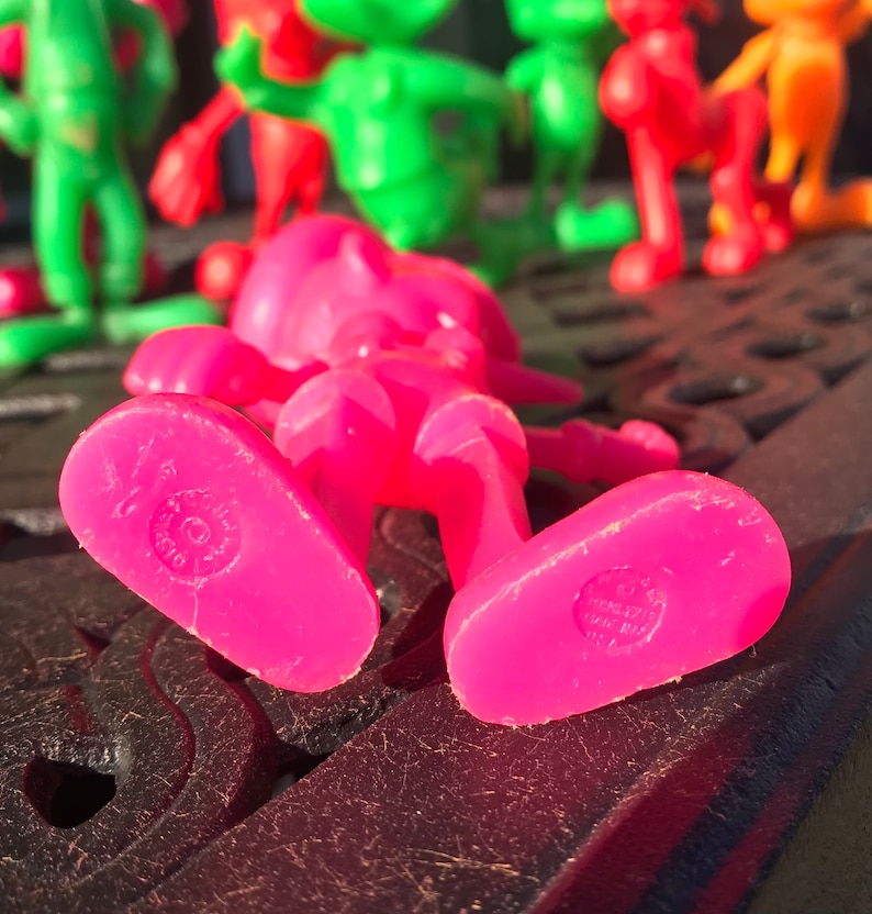 May include: A pink plastic toy figure of a person lying on its back. The figure has a simple design with no facial features. The figure is made of a bright pink plastic and has a smooth surface. The figure is lying on a black surface.