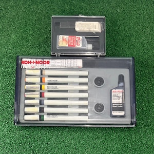 May include: A set of KOH-I-NOOR Rapidograph technical pens and accessories in a clear plastic case. The set includes pens with colored bands, black ink, and other tools. The case is on a green surface.