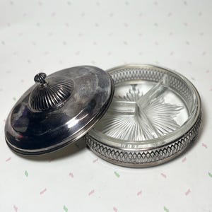 May include: A vintage silver-plated serving dish with a clear glass insert. The dish is divided into three sections and has a decorative rim. The lid is open, revealing the interior. The lid has a small, round handle.
