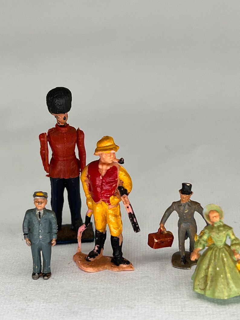 May include: A collection of vintage miniature figurines. The figures include a soldier in a red coat, a man in a yellow outfit with a pipe, a man in a suit, a man with a suitcase, and a woman in a green dress.