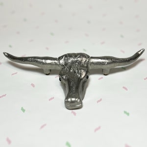 May include: A silver-colored metal sculpture of a longhorn steer head. The steer's head is detailed with textured fur and small black eyes. The horns extend outward, creating a wide span. The sculpture is a decorative item.